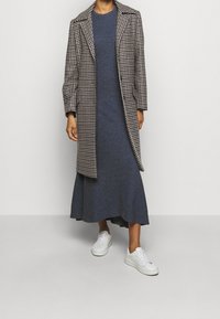 Manteau à motif pied-de-poule, robe longue bleu foncé et baskets blanches. Le manteau présente des revers crantés et des manches longues ; la robe a un ourlet évasé.