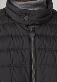 Veste noire matelassée avec un col montant, une fermeture éclair frontale à tirage doux, et des coutures horizontales discrètes sur la surface du tissu.