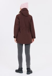 Bruine waterdichte parka met een capuchon, verstelbare taille en duimgaten in de mouwen. Draagt over een paar grijze jeans en zwarte wandelschoenen. Lichtroze beanie.