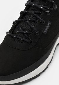 Černé semišové a síťované tenisky s kontrastními bílými šňůrkami, gumovou podrážkou a značkovým logem Timberland na boku. Šestiboké průchodky dodávají detail.