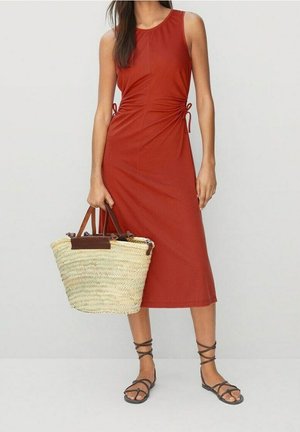 Femme portant une robe midi sans manches de couleur rouille, tenant un grand sac fourre-tout en paille tissée, et chaussée de sandales plates noires à brides.