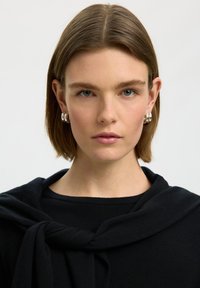 Jeune femme aux cheveux bruns courts et aux yeux bleus, portant des boucles d'oreilles anneaux en argent et un haut noir avec un pull noir posé sur ses épaules.