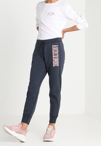 Persoon in marineblauwe joggers met "AEROBIC" tekst op het been, witte lange mouwen shirt en roze sneakers, staande met één hand op de heup.