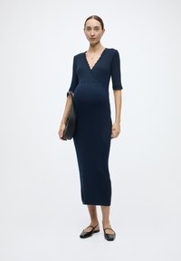 Φόρεμα από ριμπ πλεκτό ύφασμα σε ναυτικό μπλε με V-λαιμόκοψη, μανίκια με κυματιστά άκρα και εφαρμοστή σιλουέτα, συνδυασμένο με μαύρη τσάντα χειρός και μπαλαρίνες.