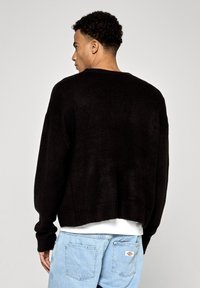 Melna adīta džemperis ar ribotām malām un piedurknēm, piedāvā relaksētu piegriezumu un kritušo plecu dizainu. Valkāts ar gaiši ziliem džinsiem.