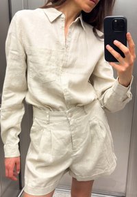 Vaalean beige-puuvilla-paita pitkillä hihoilla ja taskulla, yhdistetty puolihameisiin, joissa on poimintoja. Molemmissa vaatteissa on pehmeä, teksturoitu pinta.