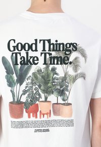 Camiseta blanca con un gráfico de plantas en macetas de colores. El texto arriba dice "Las cosas buenas llevan tiempo" en fuente negrita.