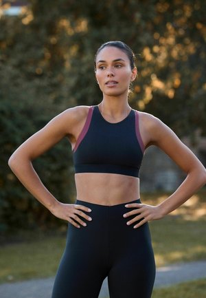 Fekete és bordó sportfelszerelésben áll egy nő a szabadban, kezeit a csípőjén tartva, oldalra néz egy elmosódott természetes háttér előtt.