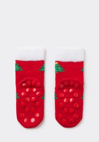 Red socks with a white fuzzy cuff, featuring green Christmas tree designs. The soles have textured red rubber dots for grip.