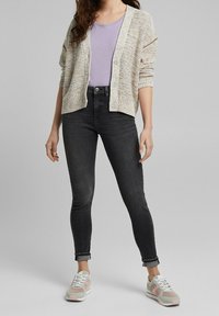 Leicht gestrickte, mehrfarbige Strickjacke über einem lavendelfarbenen Oberteil, kombiniert mit schwarzen Skinny-Jeans und grauen Sneakers mit pinken Akzenten.