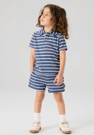 Kind mit lockigem Haar, das ein blau-weiß gestreiftes Kurzarmshirt und passende Shorts, weiße Socken und beige Schuhe trägt, schaut nach oben.