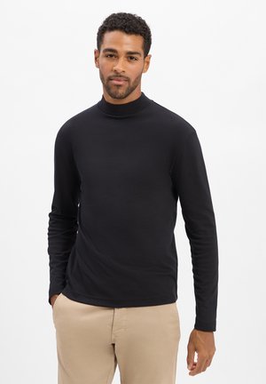 Czarna, gładka koszulka z długim rękawem i golfem, o dopasowanym kroju, noszona z beżowymi spodniami.