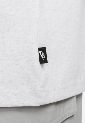 Camiseta blanca de algodón con un acabado texturizado, que presenta una etiqueta negra de Nike en la costura lateral. La camiseta tiene mangas cortas y cuello redondo.
