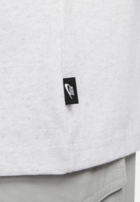 Bela tkanina v bližnjem planu, ki prikazuje majhno črno oznako Nike z belim logotipom swoosh, prišito v šiv nad svetlo sivimi hlačami.