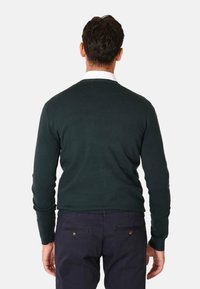 Maglione lavorato a maglia verde scuro con scollo a girocollo, maniche lunghe e polsini a costine, abbinato a chinos blu navy e dettagli visibili sulla schiena.