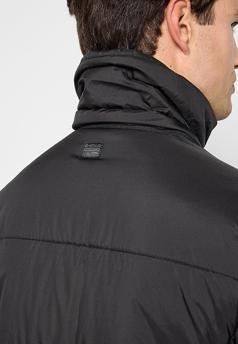 Chaqueta acolchada negra con cuello alto, textura acolchada y un parche de logo en la parte posterior, diseñada para brindarle calidez y comodidad.