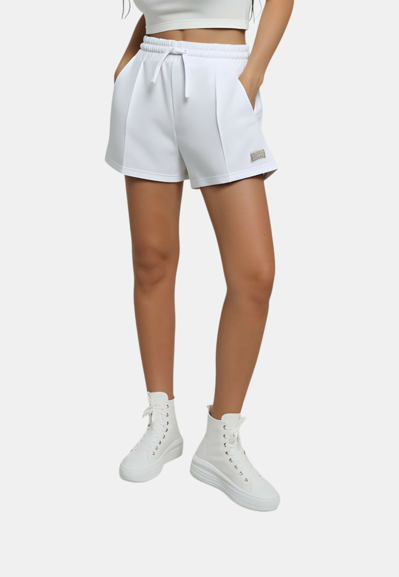 Pantaloni atletici bianchi con cinturino elastico e stringa, dotati di tasche laterali e un piccolo logo sul davanti. Abbinati a sneakers bianche alte.