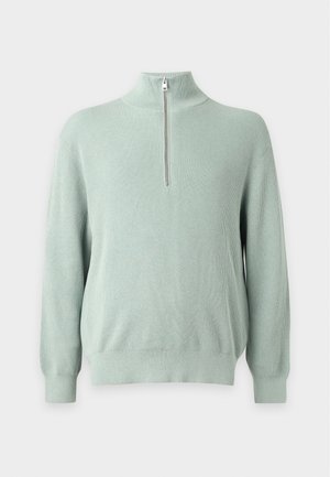 Jeune homme portant un pull vert clair à fermeture éclair au quart, un pantalon beige, des baskets blanches et un pull rayé beige et marron noué autour de sa taille.