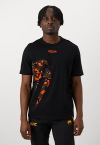 Sort t-shirt med korte ærmer, der har iøjnefaldende orange og brune camouflagegrafik på forsiden, med "VENUM"-logo i orange ved halsudskæringen.