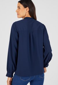 Blusa blu navy con maniche lunghe a sbuffo, colletto a volant e polsini arricciati. Presenta una piega sul retro per un maggiore movimento. Tessuto liscio.