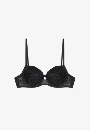 Woman with slicked-back hair wearing black lace bra and matching lace panties, standing against plain light background.