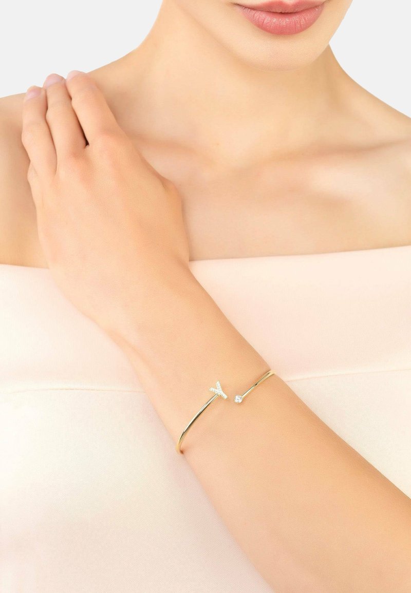 Gold bracelet featuring a delicate arrow design with small clear gemstones on one side, worn on a bare wrist against a light background.