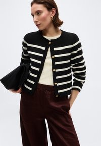 Woman wearing a black and white striped buttoned cardigan, cream top, dark brown satin pants, holding a black clutch bag.