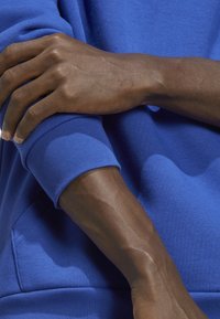 Royal blue sweateri kangasta, jossa on resorit, lähikuva kädestä, joka lepää hihassa, esitellen ihon rakennetta ja hienovaraisia yksityiskohtia.