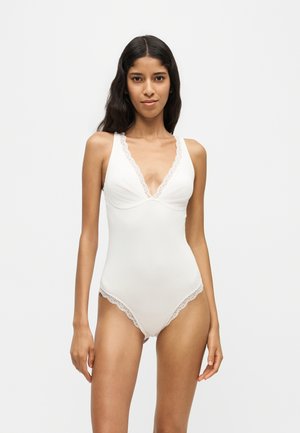 Kvinde med langt mørkt hår iført hvidt ærmeløst bodystocking med blonder, stående mod en ensfarvet lys baggrund.