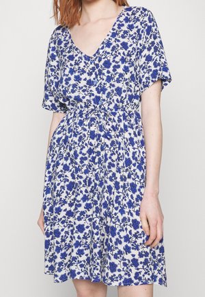Femme portant une robe blanche mi-longue avec un motif floral bleu, manches courtes, col en V et taille froncée avec des boutons sur le devant.