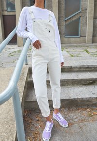 Salopette bianca in cotone con spalline regolabili, dotata di tasca e dettagli in metallo argentato, abbinata a sneaker in tela viola e una camicia bianca a maniche lunghe.