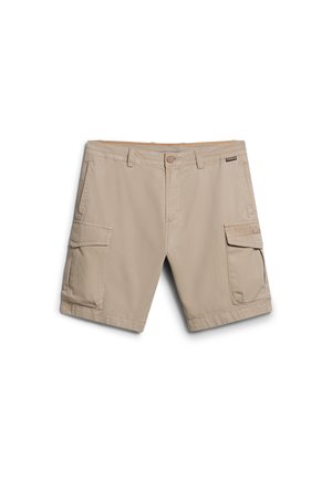 Mann trägt Khaki-Shorts, beige langärmeliges Hemd, schwarze Weste, schwarze Wanderschuhe, trägt Rucksack und hält eine grüne Wasserflasche.