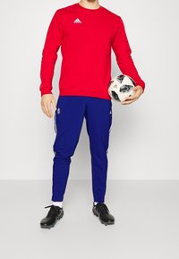 Czerwona sportowa koszula z długim rękawem i białym logo, połączona z niebieskimi spodniami dresowymi z białymi paskami. Trzyma piłkę nożną, ma na sobie czarne korki.