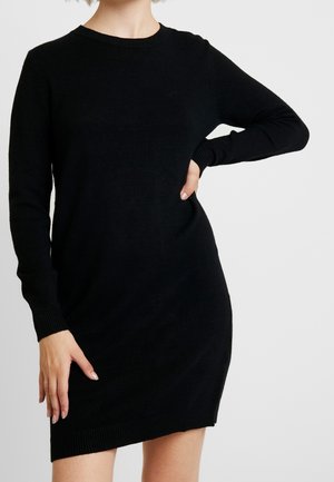 Kobieta w dopasowanej czarnej dzianinowej sukience z długim rękawem i prążkowanymi mankietami oraz dołem, pozująca z jedną ręką na biodrze na jednolitym tle.