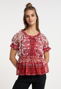 Rød blomstermønstret peplum-bluse med korte pufferede ærmer, en samlet halsudskæring og et struktureret, gennemsigtigt stof. Sorte bukser bæres nedenunder.