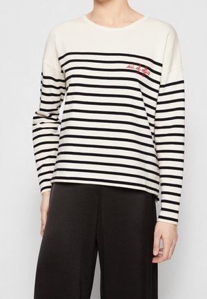 Mujer con una camisa blanca de manga larga con rayas horizontales negras y texto rojo que dice "fuera de la oficina", combinada con pantalones negros.