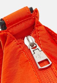 Pomarańczowa materiałowa torba z srebrnym suwakiem z logo "CK", podkreślona starannym przeszyciem i smukłym, zwężającym się designem.