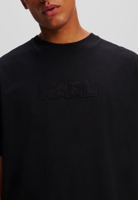 Juodas medvilninis marškinėlis su tekstūruotu siuvinėtu "KARL" logotipu, atitinkančiu juodą spalvą. Trumpomis rankovėmis ir klassikiniu apvaliomis kaklo iškirpimu.