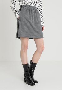 Mujer vistiendo una falda de pata de gallo en blanco y negro, una blusa de lunares y botas de charol negras de tacón medio hasta la pantorrilla, de pie frente a un fondo blanco.