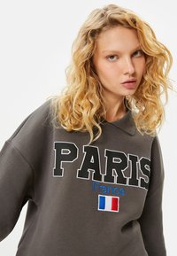 Ciemnoszara bluza z wyraźnym napisem "PARIS", niebieskim haftem "France" oraz małym akcentem w postaci francuskiej flagi. Miękki materiał, luźny krój.