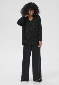 Vrouw met krullend haar in een zwarte glinsterende V-hals trui, brede zwarte broek en spitse zwarte schoenen met studs, die glimlacht en haar nek aanraakt.