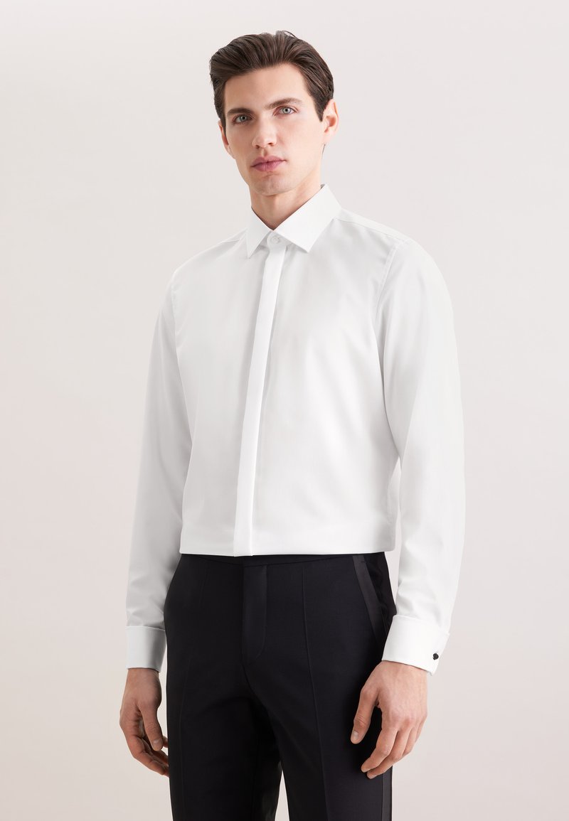 Homme portant une chemise blanche à manches longues et un pantalon noir, debout devant un fond clair uni.