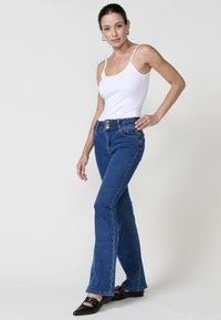 Jeans de campana azules con una cintura abotonada y bolsillos delanteros, combinados con una camiseta sin mangas blanca y zapatos planos negros. Textura de denim suave.