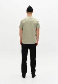 Tricou scurt cu maneci scurte de culoare verde măsliniu, cu decolteu rotund, asortat cu pantaloni negri și o caciulă neagră, văzut din spate pe un fundal simplu.