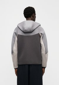 Pelēka, brūna un bēša kapuci ar sporta krekliņu; raksturo panelējums ar dažādām nokrāsām, mīksta tekstūra un brīvs piegriezums.