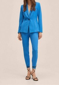 Blazer bleu sur mesure avec col cranté et fermeture à un bouton, associé à un pantalon slim. Tissu lisse, lignes épurées et détails minimalistes.