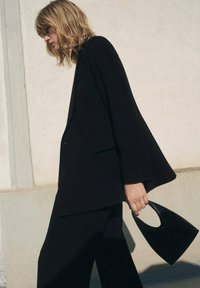 Černý strukturovaný blazer s širokými rukávy, kombinovaný s odpovídajícím outfitem. V jedné ruce drží malou zakřivenou černou kabelku. Světlé pozadí.