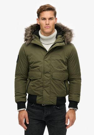Olive' zielona kurtka pikowana z kapturem wykończonym futrem, z zamkiem błyskawicznym, dwoma przednimi kieszeniami i ściągaczami przy mankietach. Noszona na lekkim swetrze.