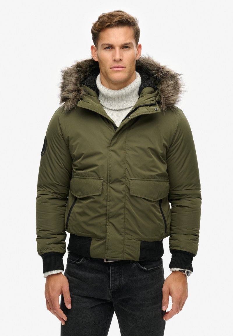 Olijfgroene gewatteerde jas met een muts met bontafwerking, ritssluiting, twee voorzakken en geribbelde manchetten. Draag het over een lichte trui.
