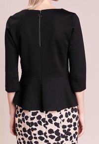 Eine Frau trägt ein schwarzes Peplum-Top mit drei Viertelärmeln und einem sichtbaren Rückenreißverschluss, kombiniert mit einem weißen Rock mit schwarzen Flecken.
